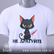 Футболка не дратуйте, білого кольору, зображена зверху, на фоні світло-сірого кольору, принт з чорним котом з червоним ножем, текст під котом чорного кольору, на футболці є етикетка з зеленим шнурком.