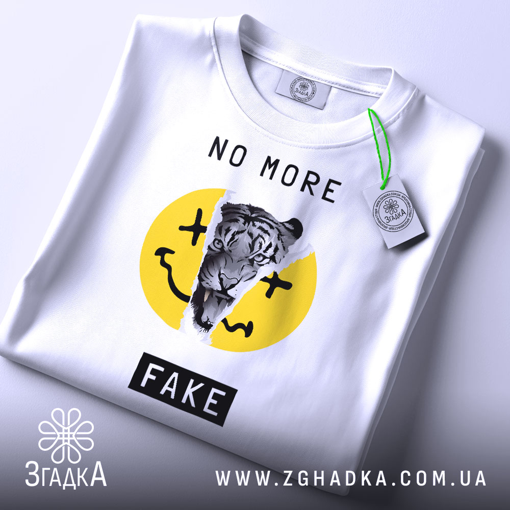 Футболка не фейк. Біла футболка, складена на столі, з принтом тигра на жовтому фоні, текстом 'NO MORE FAKE' над принтом і чорним написом 'FAKE' внизу. На футболці є етикетка з зеленим шнурком. Фон світло-сірий.