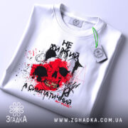 Футболка не хижий, біла, зображена зверху, складена на поверхні, фон світло-сірий, принт з черепом, червоними плямами та чорними елементами, з етикеткою.