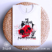 Футболка не хижий, біла, зображена зверху, складена на плетеній кошику, фон білий, принт з черепом, червоними плямами та чорними елементами.