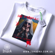 Футболка не мовчи подарунок, біла, складена на поверхні, фон світло-сірий, принт з зображенням жінки в чорному одязі з червоними плямами, написом 'НЕ МОВЧИ' і українським прапором на фоні, з етикеткою.