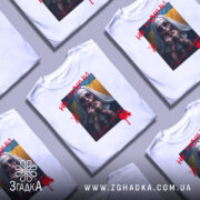 Футболка не мовчи подарунок, біла, розташована на поверхні разом з іншими футболками, фон світло-сірий, принт з зображенням жінки в чорному одязі з червоними плямами, написом 'НЕ МОВЧИ' і українським прапором на фоні.