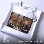 Футболка не нам судити. Біла футболка, складена на столі, з принтом трьох персонажів з анімації, які обговорюють щось. На футболці є етикетка з зеленим шнурком.