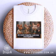 Футболка не нам судити. Біла футболка, складена на плетеному кошику, з принтом трьох персонажів з анімації, які спілкуються. На фоні видно текст на футболці.