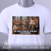 Футболка не нам судити. Біла футболка, складена на столі, з принтом трьох персонажів з анімації, які обговорюють щось. На футболці є етикетка з зеленим шнурком.