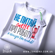 Футболка не питай мене, біла, з коротким рукавом, на столі, фон світло-сірий, принт з текстом червоного та блакитного кольорів, з малюнком олівця, що кричить, з етикеткою на шнурку.