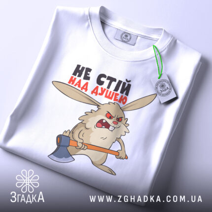 Футболка не стій над душею, біла, складена на поверхні, фон світло-сірий, принт з кроликом з сокирою, текст чорного та червоного кольору, на футболці є етикетка з зеленим шнурком.