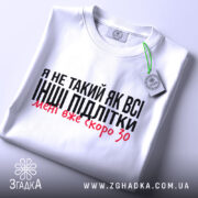 Футболка не такий як всі, білого кольору, складена на столі з верхнього ракурсу, фон світло-сірий, принт чорного кольору з червоними акцентами, на футболці є етикетка з зеленим шнурком.