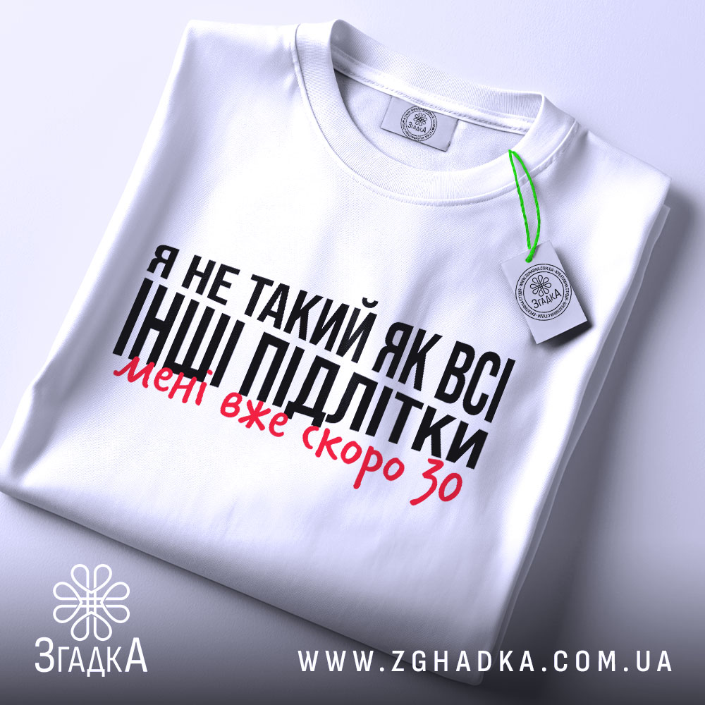 Футболка не такий як всі, білого кольору, складена на столі з верхнього ракурсу, фон світло-сірий, принт чорного кольору з червоними акцентами, на футболці є етикетка з зеленим шнурком.