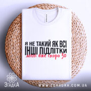 Футболка не такий як всі, білого кольору, складена на плетеній основі з верхнього ракурсу, фон світло-сірий, принт чорного кольору з червоними акцентами, текст говорить про те, що людина не така як інші підлітки.