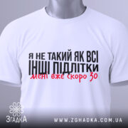 Футболка не такий як всі, білого кольору, складена на столі з верхнього ракурсу, фон світло-сірий, принт чорного кольору з червоними акцентами, на футболці є етикетка з зеленим шнурком.