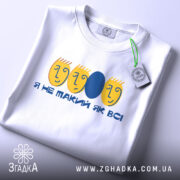 Футболка не такий як всі, біла, зображена в складеному вигляді, фон світло-сірий, принт з жовтими обличчями та синім овалом, текст "Я НЕ ТАКИЙ ЯК ВСІ", з етикеткою на зеленому шнурку.