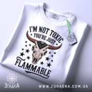 Футболка не токсичний, біла, зображена зверху, складена, фон світло-сірий, принт з черепом з рогами та текстом 'I'M NOT TOXIC YOU'RE JUST FLAMMABLE' у чорному та червоному кольорах, на ярлику зелена стрічка.