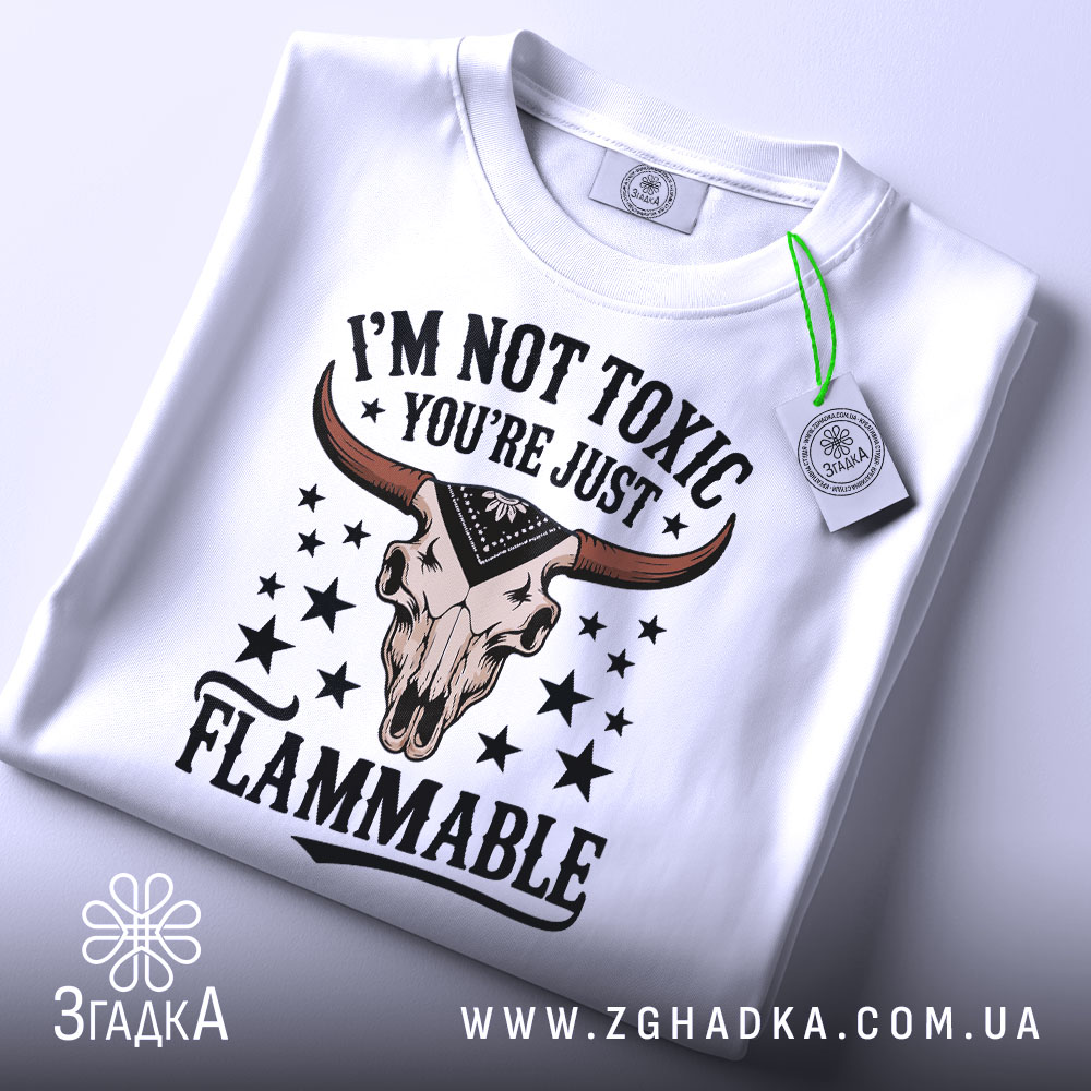 Футболка не токсичний, біла, зображена зверху, складена, фон світло-сірий, принт з черепом з рогами та текстом 'I'M NOT TOXIC YOU'RE JUST FLAMMABLE' у чорному та червоному кольорах, на ярлику зелена стрічка.