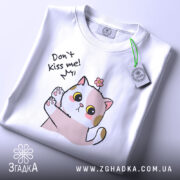 Футболка не цілуй, біла, складена на поверхні, фон світлий, принт з котиком, що піднімає лапку, з текстом 'Don't kiss me!' та етикеткою з зеленим шнурком.