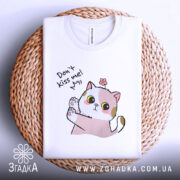 Футболка не цілуй, біла, складена на кошику, фон світлий, принт з котиком, що піднімає лапку, з текстом 'Don't kiss me!' та квіткою на голові.