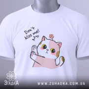 Футболка не цілуй, біла, складена на поверхні, фон світлий, принт з котиком, що піднімає лапку, з текстом 'Don't kiss me!' та етикеткою з зеленим шнурком.