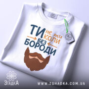 Футболка не ти без бороди. Біла футболка, складена на столі, з видимим етикеткою. Принт з текстом 'ТИ не ти КОЛИ БЕЗ БОРОДИ' та бородою, розташований на передній частині. Фон світлий, що підкреслює футболку.
