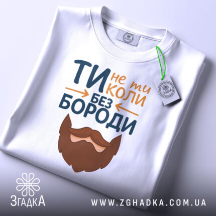Футболка не ти без бороди. Біла футболка, складена на столі, з видимим етикеткою. Принт з текстом 'ТИ не ти КОЛИ БЕЗ БОРОДИ' та бородою, розташований на передній частині. Фон світлий, що підкреслює футболку.