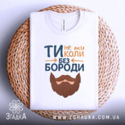 Футболка не ти без бороди. Біла футболка, складена на плетеному кошику, з текстом 'ТИ не ти КОЛИ БЕЗ БОРОДИ' та малюнком бороди. Фон світлий, що підкреслює текст і принт.