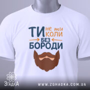 Футболка не ти без бороди. Біла футболка, складена на столі, з видимим етикеткою. Принт з текстом 'ТИ не ти КОЛИ БЕЗ БОРОДИ' та бородою, розташований на передній частині. Фон світлий, що підкреслює футболку.