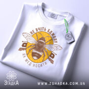 Футболка не вчіть бджолу, біла, складена на поверхні, зображена з верхнього ракурсу, фон світлий, принт з бджолою та написом у коричневих тонах на жовтому фоні, з етикеткою зеленого кольору.