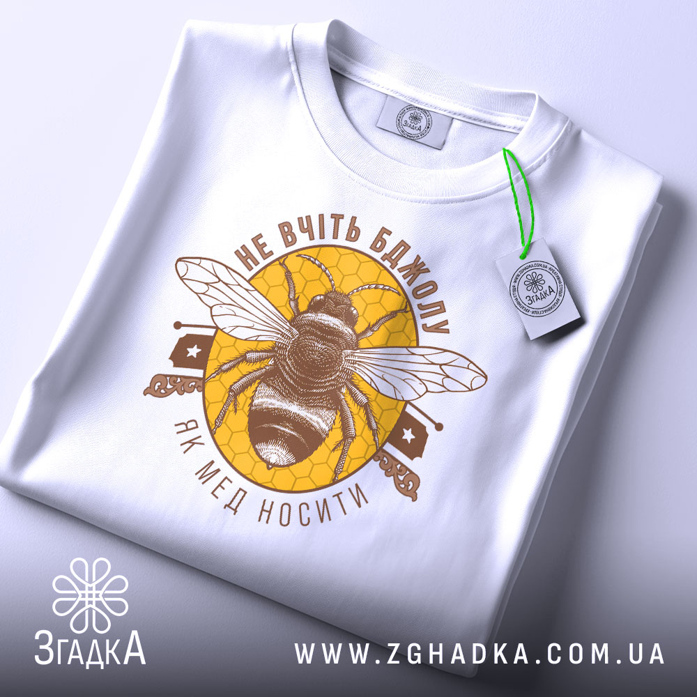 Футболка не вчіть бджолу, біла, складена на поверхні, зображена з верхнього ракурсу, фон світлий, принт з бджолою та написом у коричневих тонах на жовтому фоні, з етикеткою зеленого кольору.