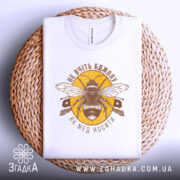 Футболка не вчіть бджолу, біла, складена на плетеній основі, зображена з верхнього ракурсу, фон світлий, принт з бджолою та написом у коричневих тонах на жовтому фоні.