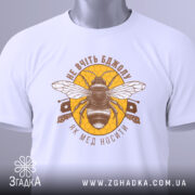 Футболка не вчіть бджолу, біла, складена на поверхні, зображена з верхнього ракурсу, фон світлий, принт з бджолою та написом у коричневих тонах на жовтому фоні, з етикеткою зеленого кольору.