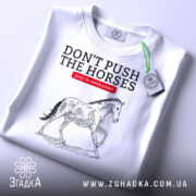 Футболка не жени коней, біла, зображена на столі, фон світлий, принт з конем у стилі малюнка, текст червоний на чорному.