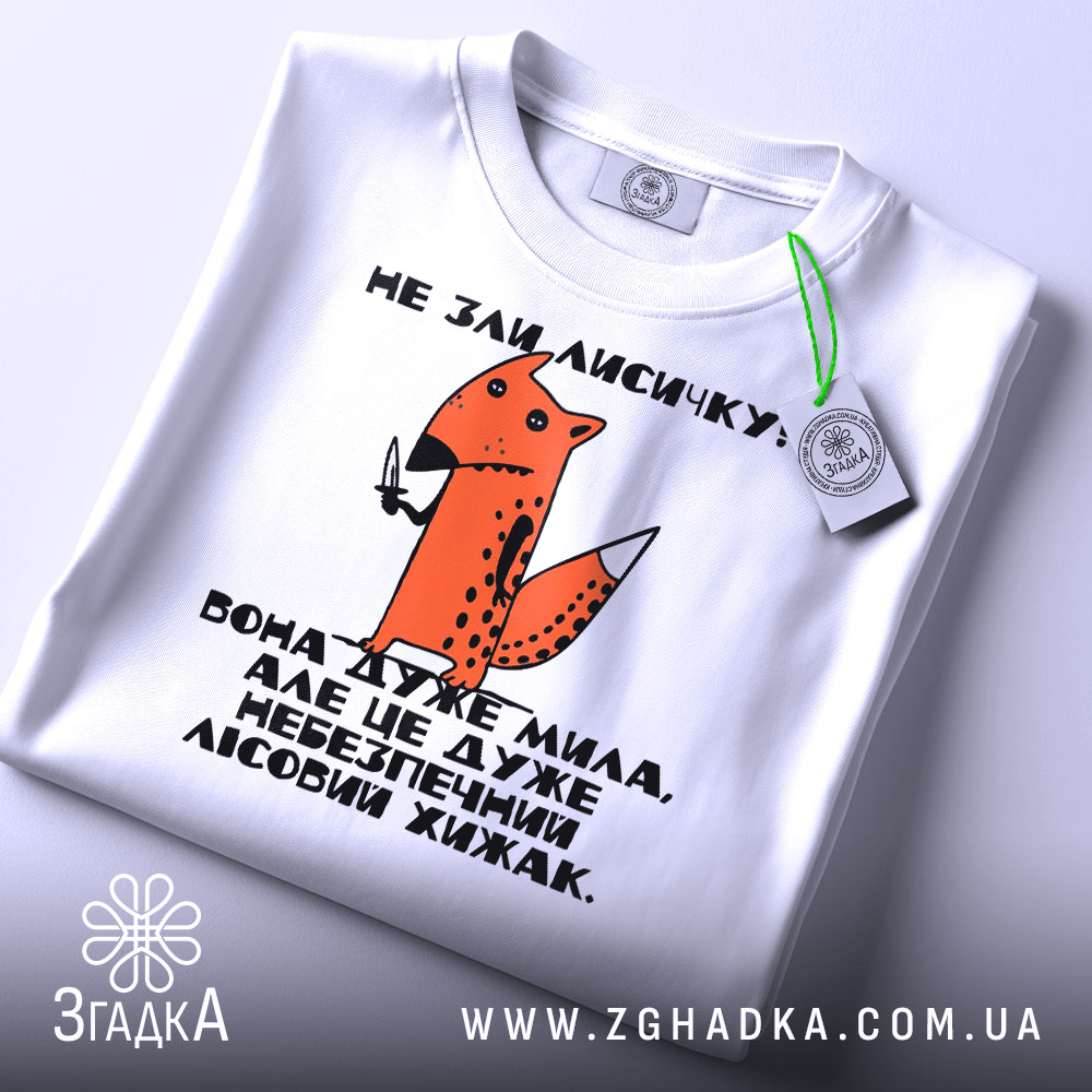 Футболка не зли лисичку. Біла футболка, складена на столі, принт з лисичкою та текстом, зелена етикетка з логотипом на комірці, фон світло-сірий.