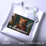 Футболка не зли лисичку. Біла футболка, складена на поверхні, принт з лисичкою з бензопилами, зелений тег на комірці, фон світлий.