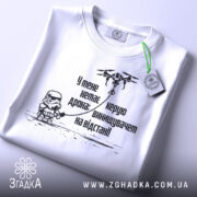 Футболка немає дрона. Біла футболка, складена на столі, з видимим етикеткою, фон світло-сірий. Принт чорного кольору з малюнком персонажа в шоломі та дрона, текст українською мовою.