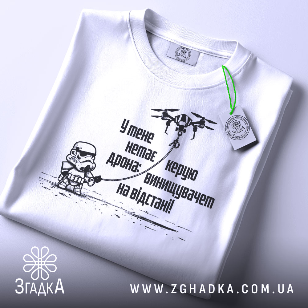 Футболка немає дрона. Біла футболка, складена на столі, з видимим етикеткою, фон світло-сірий. Принт чорного кольору з малюнком персонажа в шоломі та дрона, текст українською мовою.