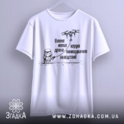 Футболка немає дрона. Біла футболка, підвішена на вішалці, фон світло-сірий. Принт чорного кольору з малюнком персонажа в шоломі та дрона, текст українською мовою.