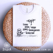 Футболка немає дрона. Біла футболка, складена на плетеному кошику, фон білий. Принт чорного кольору з малюнком персонажа в шоломі та дрона, текст українською мовою.