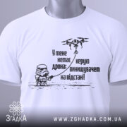 Футболка немає дрона. Біла футболка, складена на столі, з видимим етикеткою, фон світло-сірий. Принт чорного кольору з малюнком персонажа в шоломі та дрона, текст українською мовою.