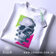 Футболка неоновий череп, біла, складена на поверхні, фон світло-сірий, принт з черепом, розділеним на смуги, з яскравими зеленими та рожевими геометричними формами, з етикеткою на зеленому шнурку.
