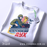 Футболка непереможний дух, білого кольору, складена на столі, фон світло-сірий, принт з військовим, що тримає автомат і піднятою рукою, на фоні червоні будівлі та дрони, з зеленим ярликом.
