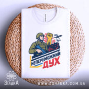 Футболка непереможний дух, білого кольору, складена на плетеній основі, фон білий, принт з військовим, що тримає автомат і піднятою рукою, на фоні червоні будівлі та дрони.