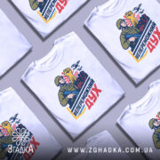 Футболка непереможний дух, білого кольору, зображена на манекені з правого боку, фон світло-сірий, принт з військовим, що тримає автомат і піднятою рукою, на фоні червоні будівлі та дрони.