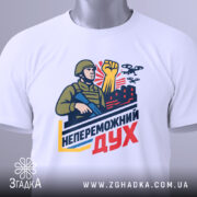 Футболка непереможний дух, білого кольору, складена на столі, фон світло-сірий, принт з військовим, що тримає автомат і піднятою рукою, на фоні червоні будівлі та дрони, з зеленим ярликом.