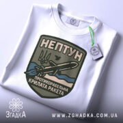 Футболка нептун крилата ракета, біла, зображена зверху, на ній прикріплений тег, фон світло-сірий, принт з зображенням ракети та текстом на темно-зеленому фоні.