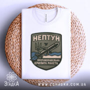 Футболка нептун крилата ракета, біла, зображена зверху на плетеній кошику, фон білий, принт з зображенням ракети та текстом на темно-зеленому фоні.
