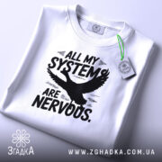 Футболка нервові системи білого кольору, складена на столі, фон світло-сірий, принт чорного кольору з написом 'ALL MY SYSTEMS ARE NERVOUS.' та зображенням орла, на комірці є етикетка.