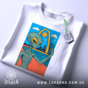 Футболка неземний вайб, біла, складена на столі, фон світло-сірий, принт з інопланетянином, що робить селфі, з етикеткою на рукаві.