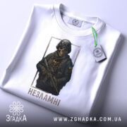 Футболка незламні воїни, біла, зображена в горизонтальному положенні, на фоні світло-сірого градієнту, принт з зображенням воїна в масці та бронежилеті, з написом 'Незламні' під ним, з етикеткою на шнурку.