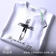 Футболка незламність духу, білого кольору, складена на столі, фон світлий, принт чорного кольору з зображенням хреста та написом 'Незламність духу', на комірці видна етикетка.