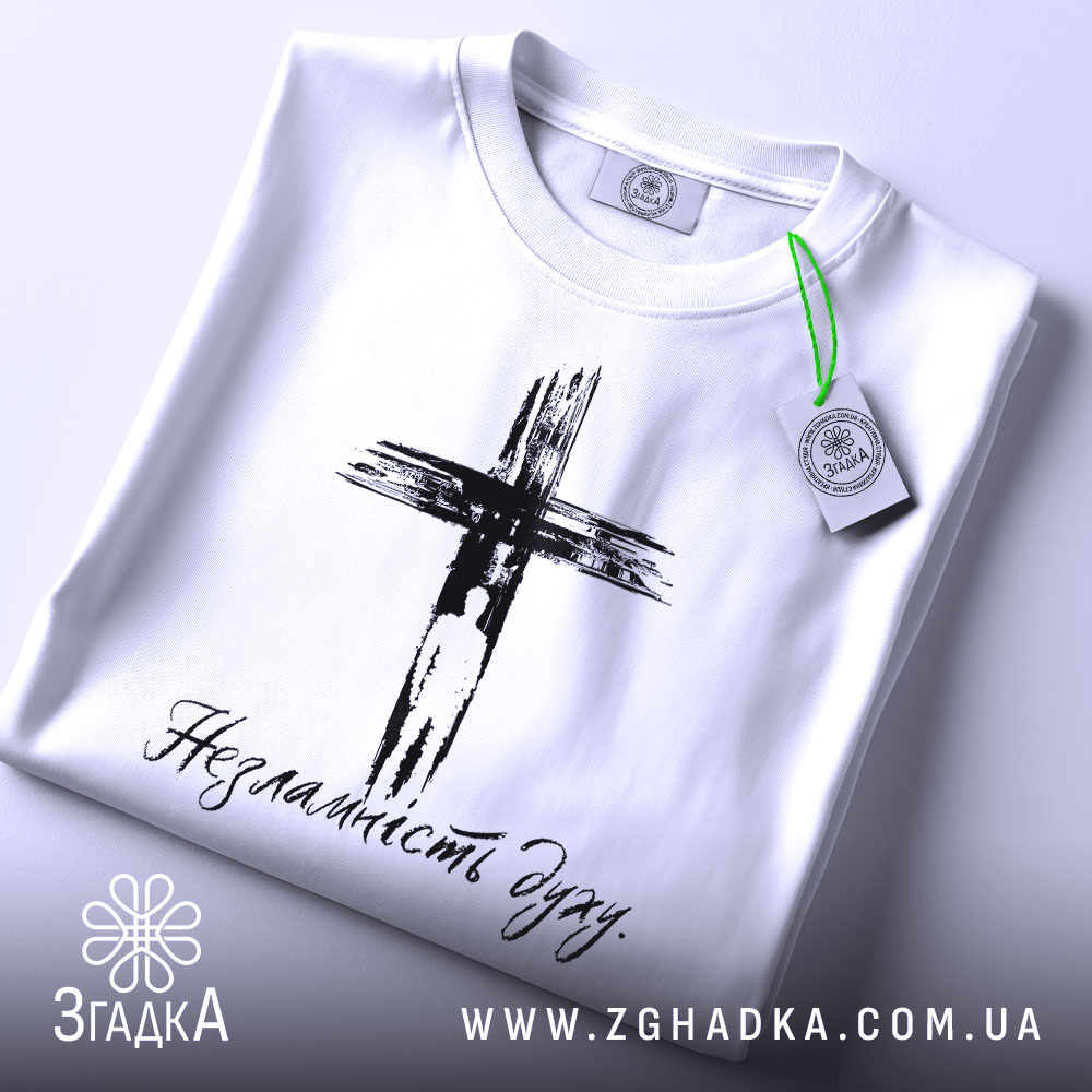 Футболка незламність духу, білого кольору, складена на столі, фон світлий, принт чорного кольору з зображенням хреста та написом 'Незламність духу', на комірці видна етикетка.
