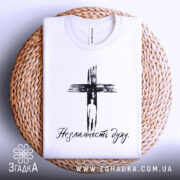 Футболка незламність духу, білого кольору, складена на плетеній основі, фон світлий, принт чорного кольору з зображенням хреста та написом 'Незламність духу'.